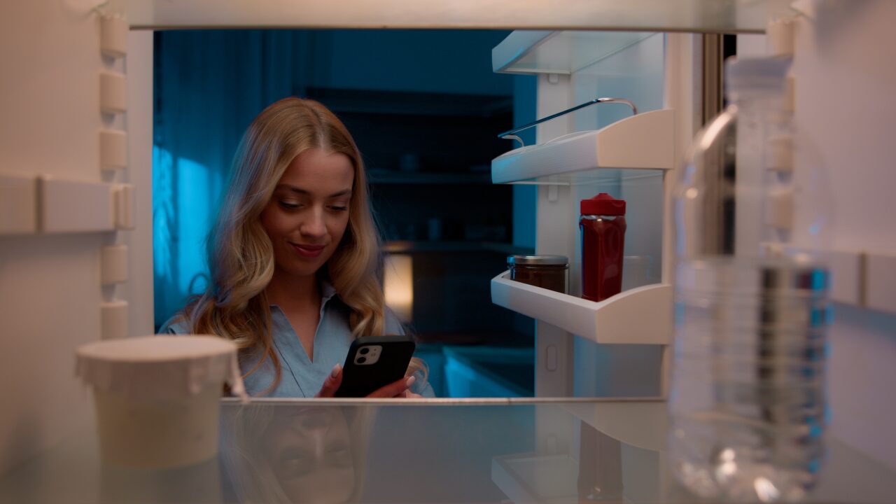 Kobieta patrząca na telefon stojąc przed otwartą lodówką.