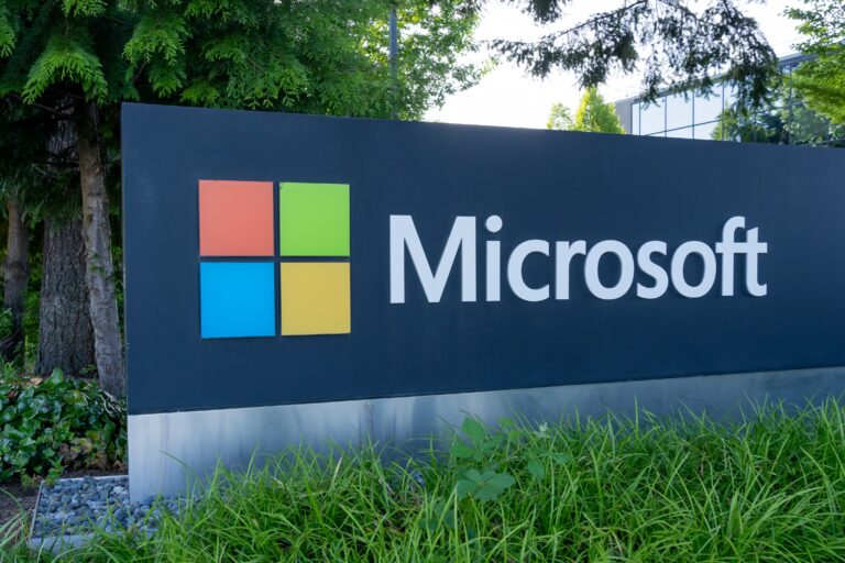 Logo Microsoft na tablicy przed budynkiem otoczonym roślinnością.