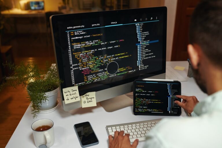 Osoba siedząca przy biurku, pisząca kod na komputerze i tablecie, otoczona rośliną doniczkową, kubkiem herbaty i telefonem komórkowym.