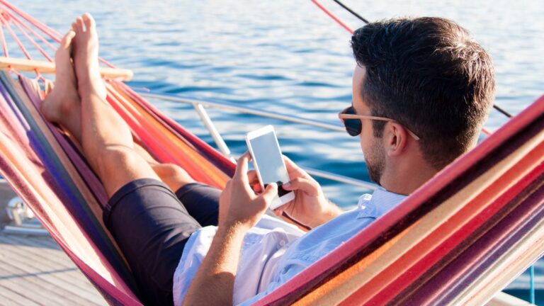 Mężczyzna w okularach przeciwsłonecznych leży w kolorowym hamaku na łódce, trzymając smartfona nad wodą.