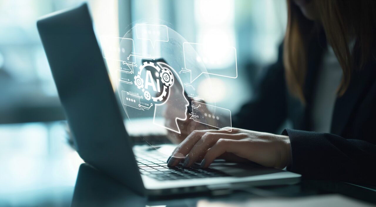 Osoba pracująca na laptopie, z holograficznym obrazem głowy robota ze słowem "AI" i ikonami technologii.