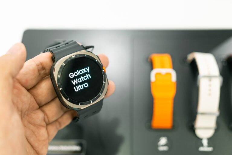 Smartwatch Galaxy Watch Ultra trzymany w ręce, na jasnym tle z dwoma paskami do zegarka w tle.