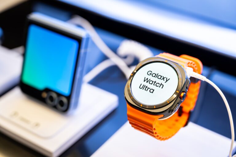 Smartwatch Galaxy Watch Ultra z jasnopomarańczowym paskiem na wystawie, w tle rozmyty telefon komórkowy.