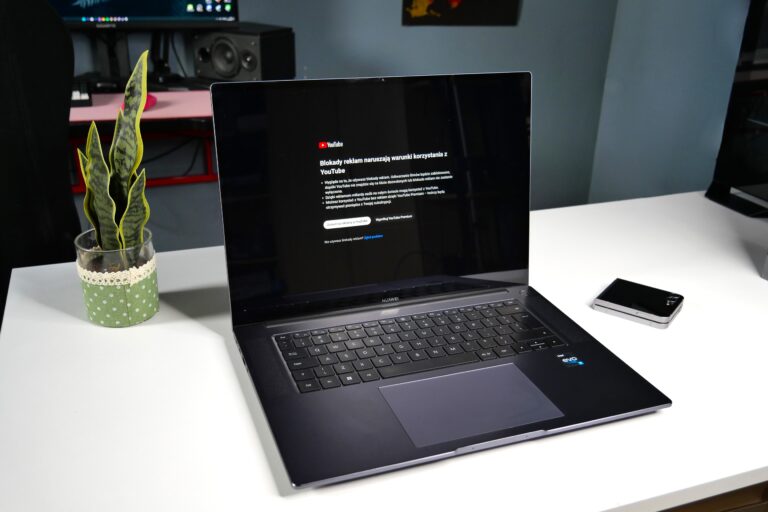Laptop na biurku z komunikatem na ekranie o blokadzie reklam na YouTube, obok mała roślina w doniczce i smartfon.