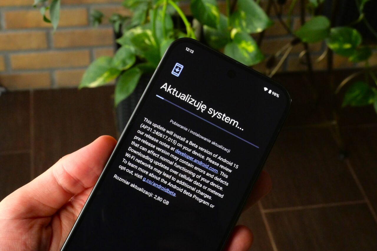 Smartfon z ekranem wyświetlającym aktualizację systemu Android, trzymany w ręku na tle roślin.