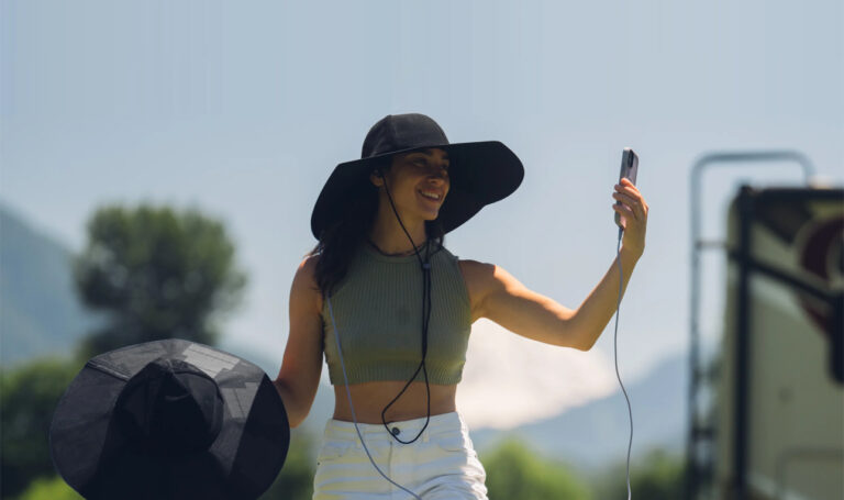 Kobieta w kapeluszu robiąca selfie na zewnątrz, trzymając w ręku słuchawki.