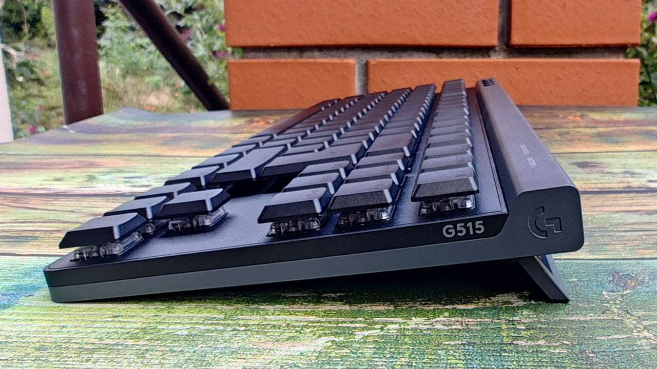 Czarna klawiatura mechaniczna Logitech G515 z widocznym logo stoi na szorstkim, zielonym stole na tle ceglanej ściany.