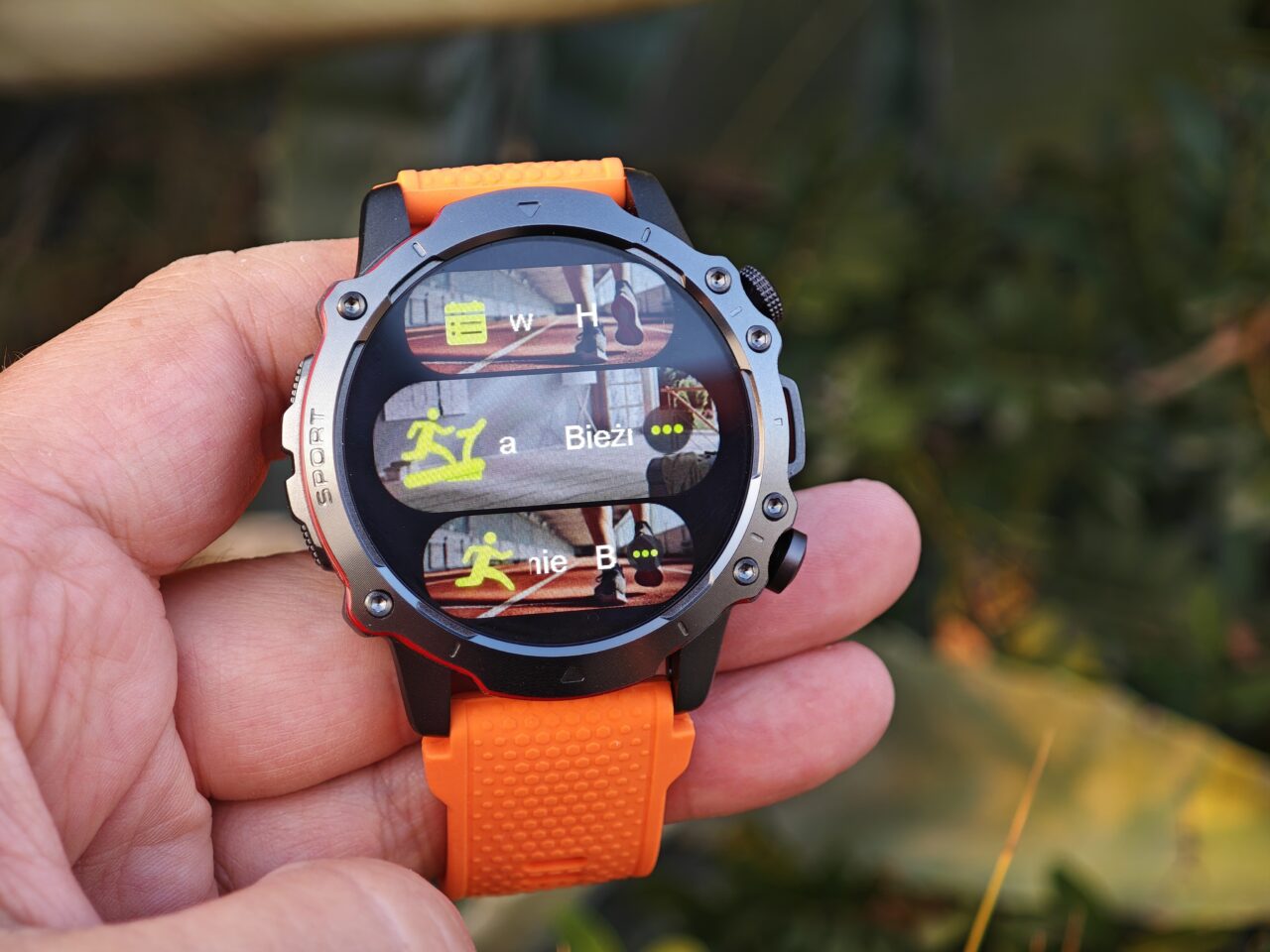 Smartwatch z pomarańczowym paskiem, wyświetlający ekran wyboru trybów ćwiczeń: bieżnia, bieganie.