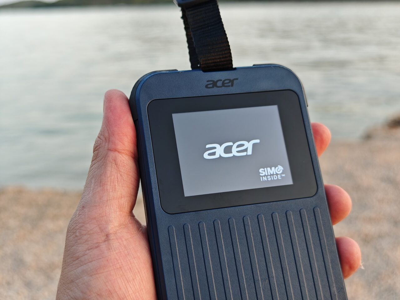 Osoba trzymająca urządzenie Acer Connect Enduro 3 z wyświetlaczem, na którym widoczne jest logo Acer i napis "SIMO INSIDE", w tle widać wodę i wybrzeże.