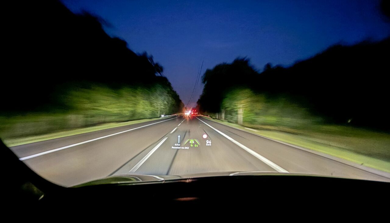 Widok z wnętrza samochodu na autostradę w nocy z wyświetlaną na szybie prędkością i odległością do obiektu.