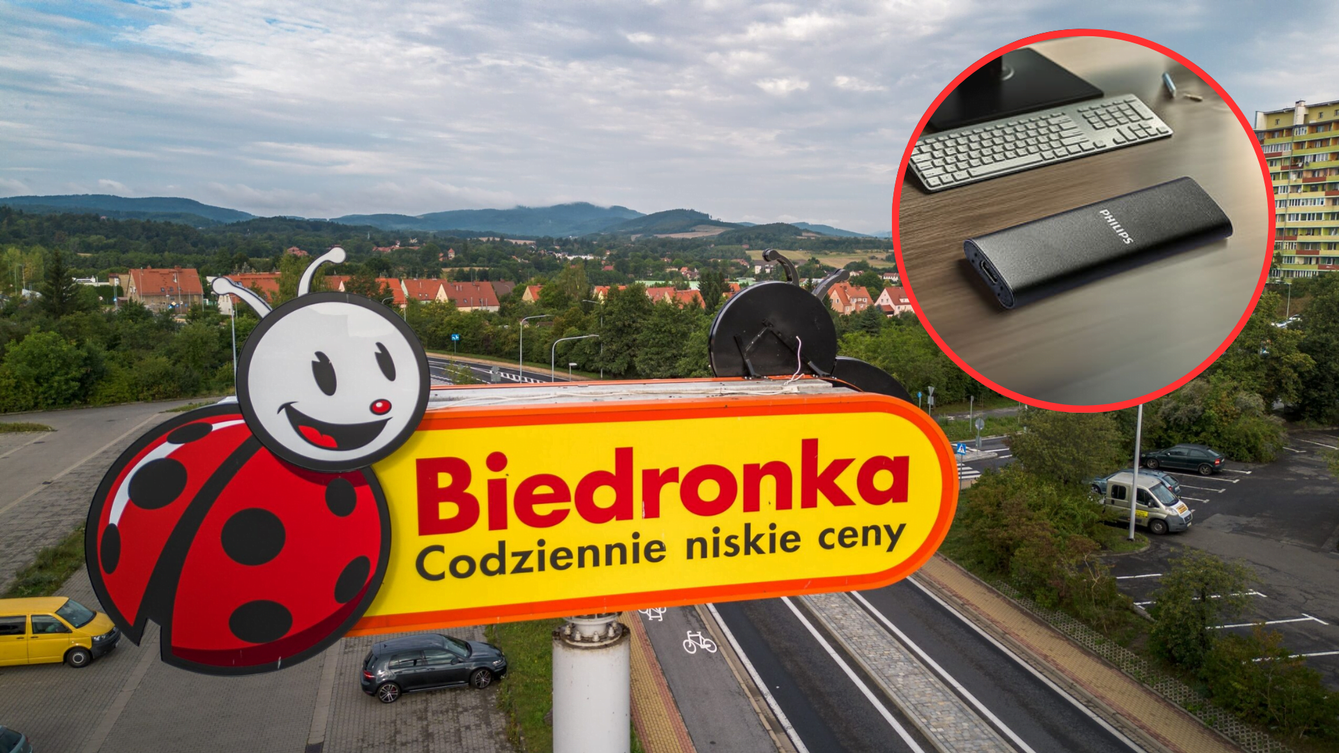 Szyld sklepu Biedronka z napisem "Codziennie niskie ceny" na tle osiedla i gór, w rogu zdjęcia doda owal z klawiaturą komputerową i powerbankiem marki Philips na biurku.