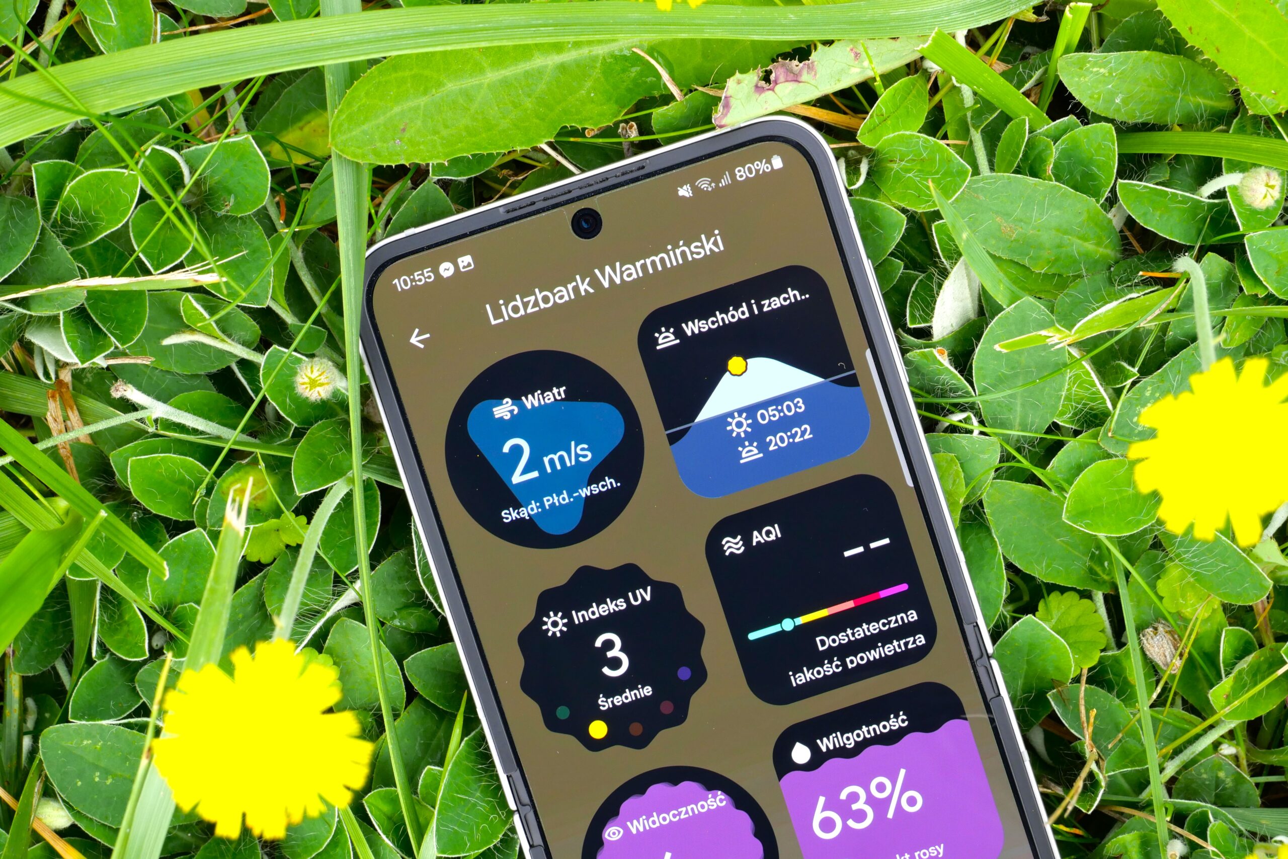 Smartfon wyświetlający dane pogodowe dla Lidzbarka Warmińskiego na tle trawy.