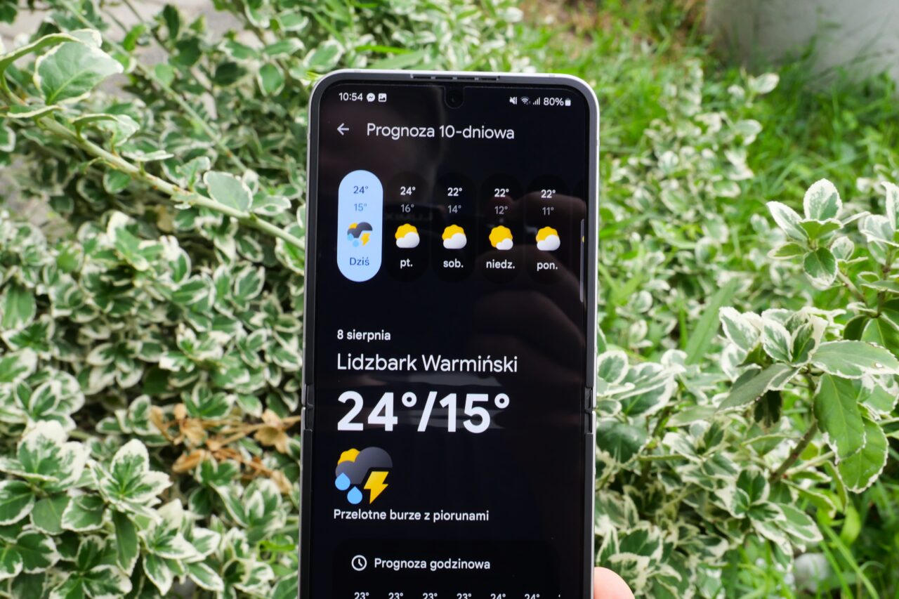Ekran telefonu z prognozą pogody na 10 dni dla Lidzbarka Warmińskiego na tle zielonych krzewów.