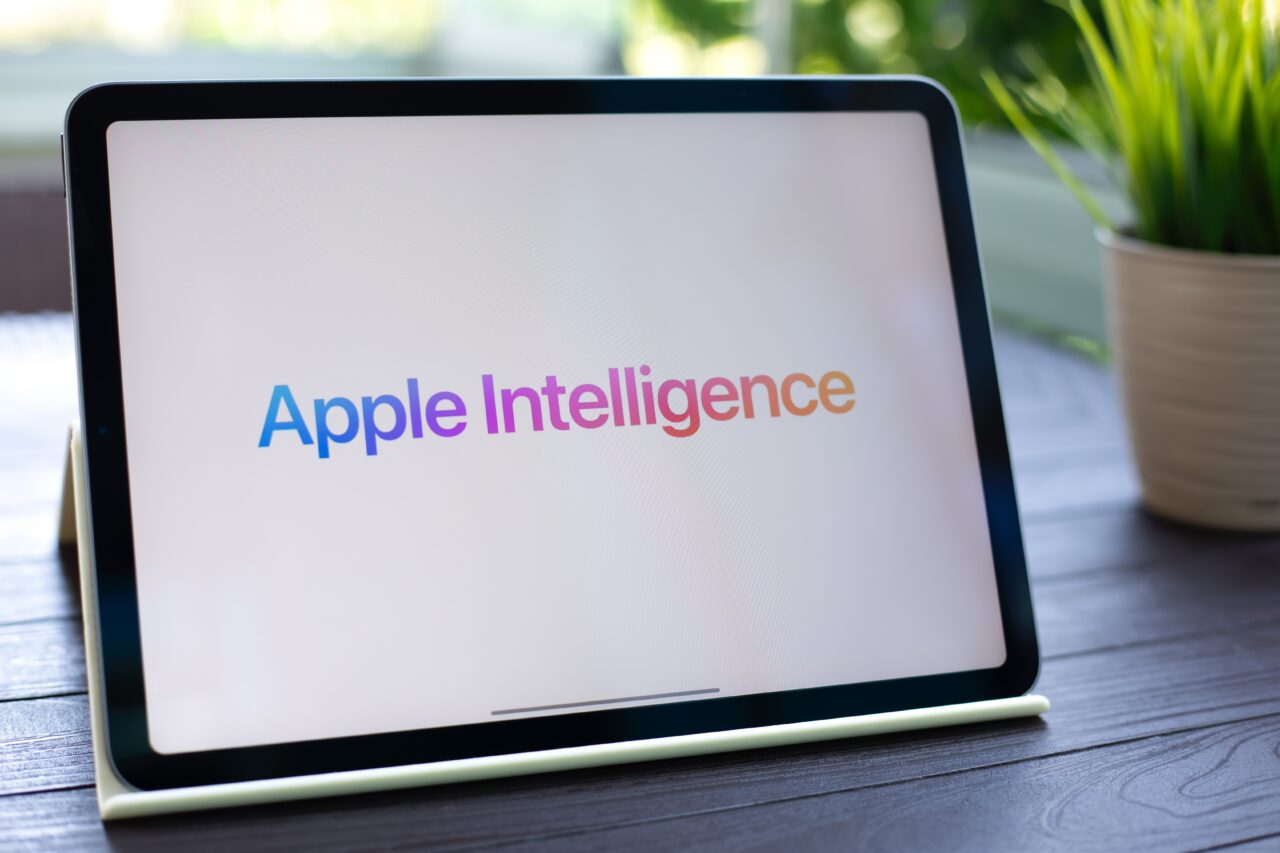 iOS 18.1 Tablet z napisem "Apple Intelligence" na ekranie, stojący na biurku obok doniczki z rośliną. 