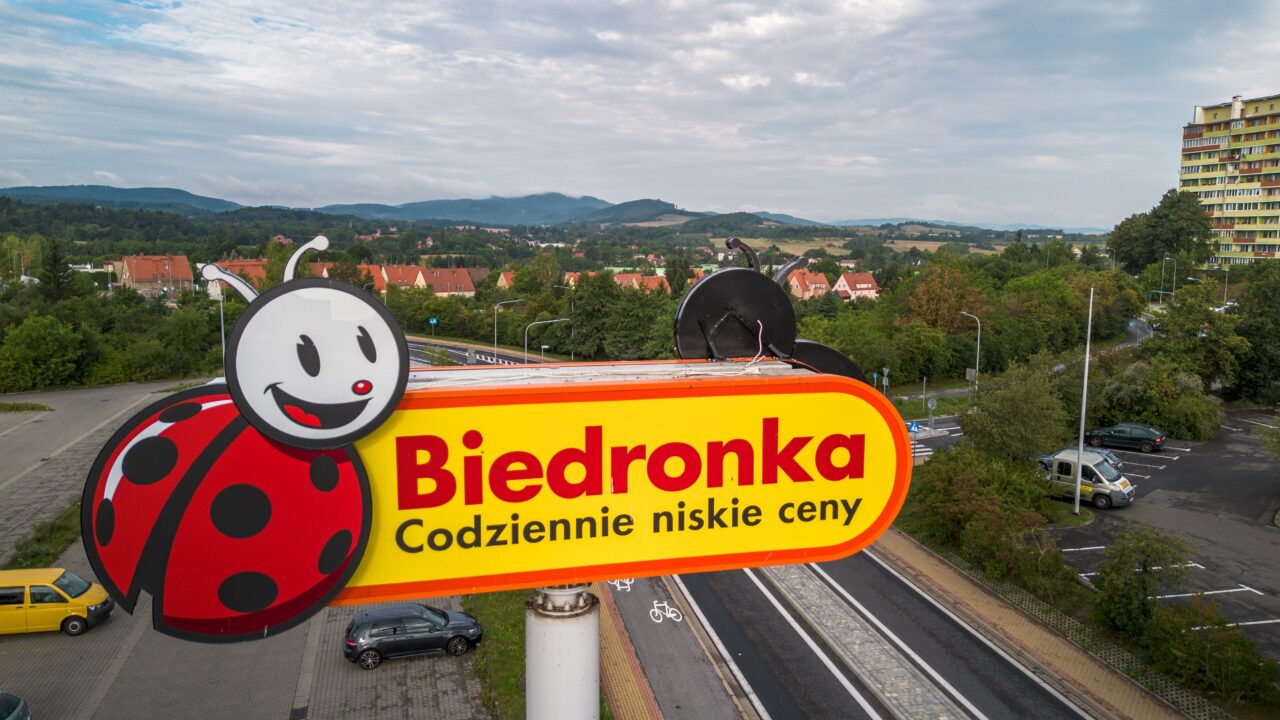 Szyld sklepu Biedronka z hasłem "Codziennie niskie ceny" na tle krajobrazu z budynkami i zielenią.