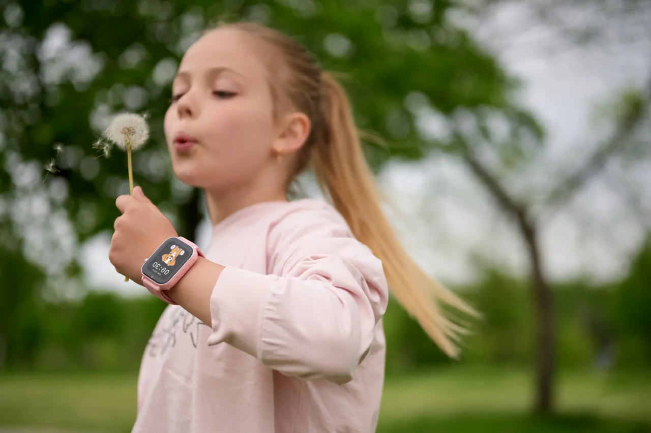 Dziewczynka dmuchająca na dmuchawca, z różowym smartwatchem na nadgarstku. Lokalizator dla dziecka jej pomoże