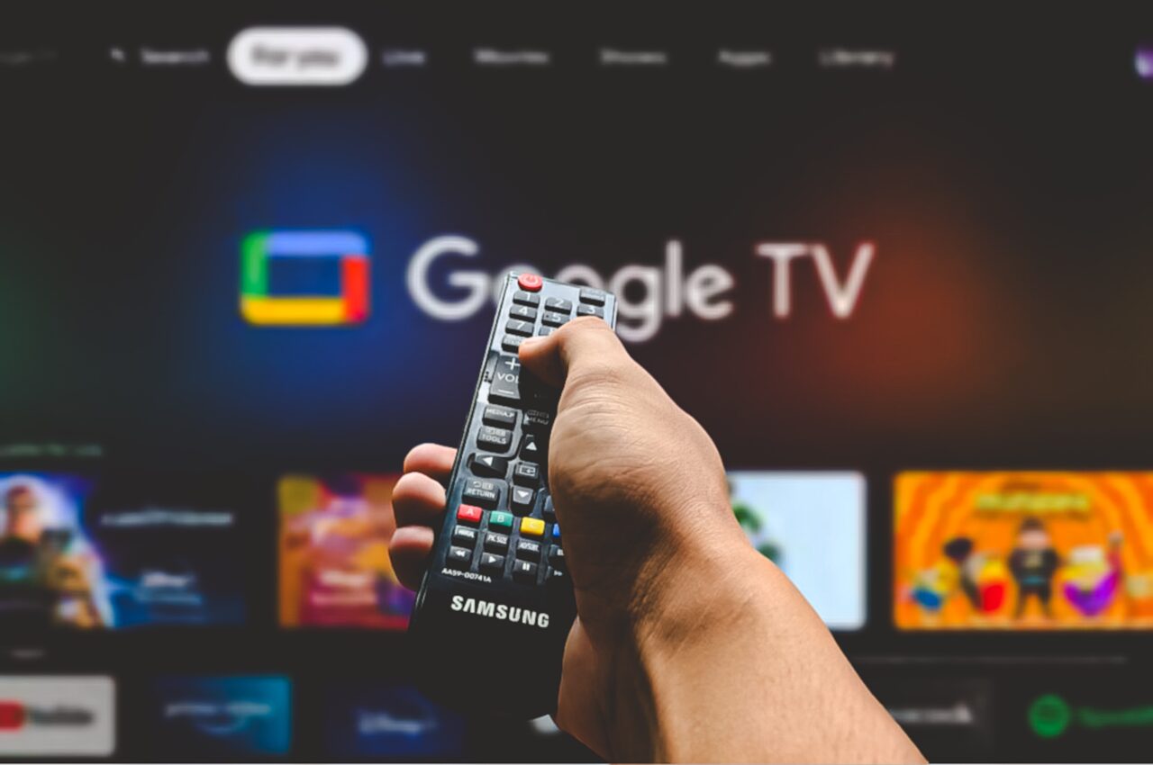 Ręka trzymająca pilot Samsung, na tle ekranu z interfejsem Google TV.