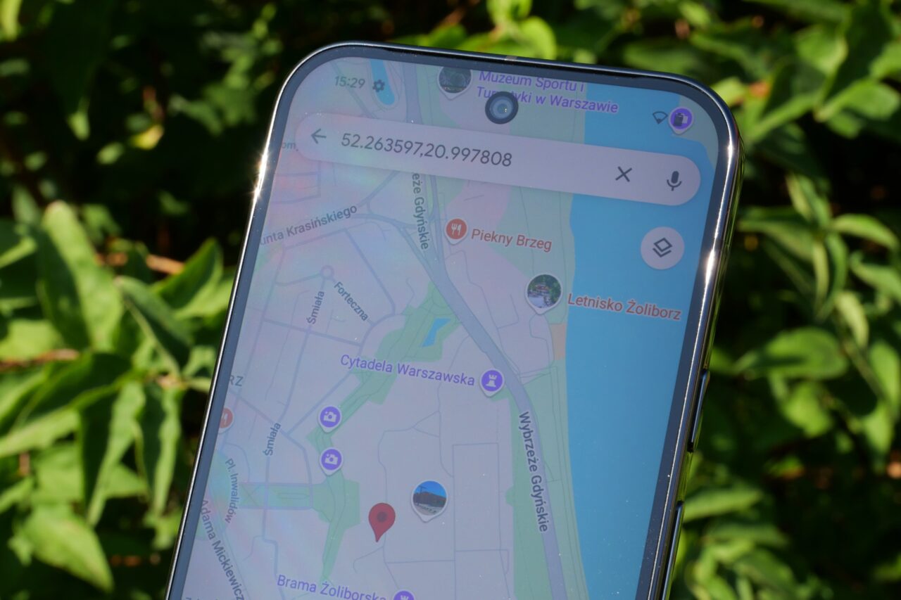 Smartfon z wyświetloną mapą Google Maps i współrzędnymi GPS, w tle liście drzew.