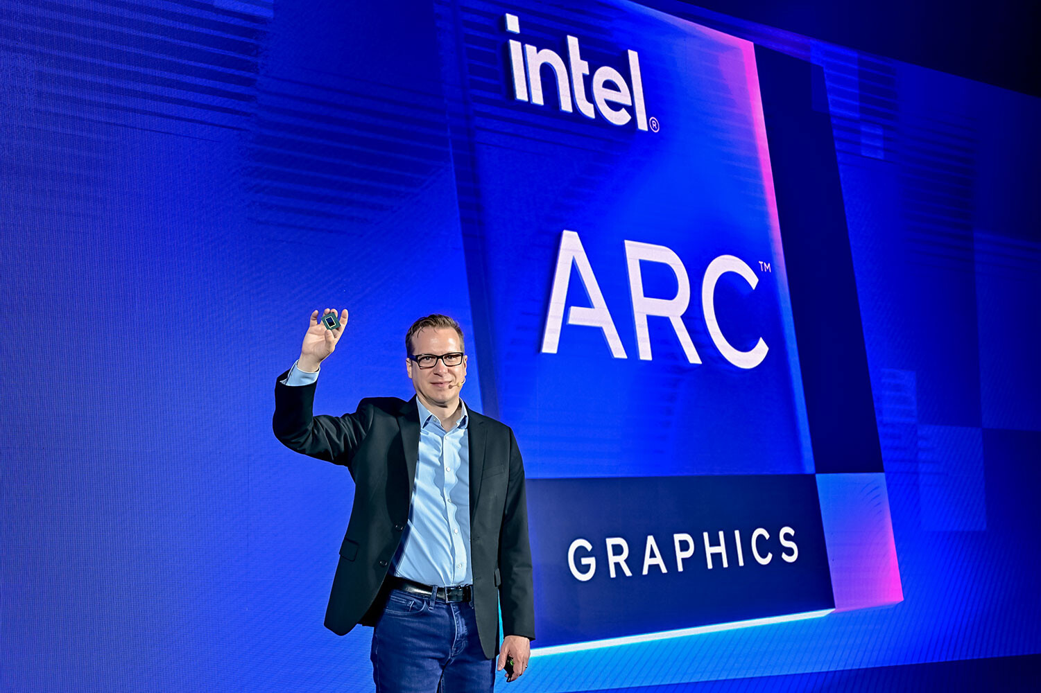 Mężczyzna trzymający chip Intel Arc A760A przed dużym ekranem z napisem "Intel ARC Graphics".