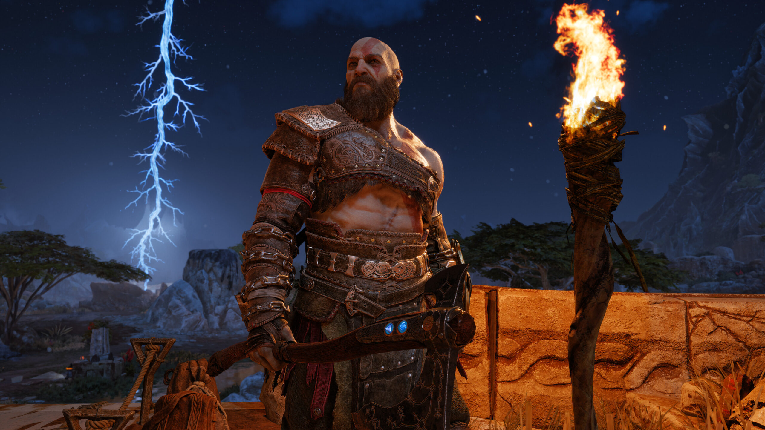 God of War Ragnarok PC. Brodaty wojownik z toporem w ręku, ubrany w zbroję, stoi obok płonącej pochodni na tle krajobrazu z błyskawicą na nocnym niebie.