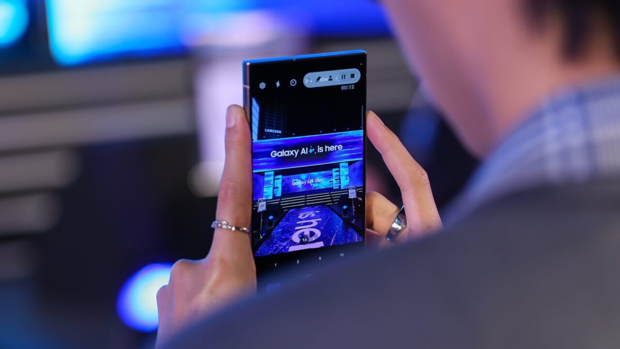  one ui 6.1.1 Osoba robiąca zdjęcie telefonu komórkowego wyświetlającego napis "Galaxy AI is here" podczas wydarzenia.