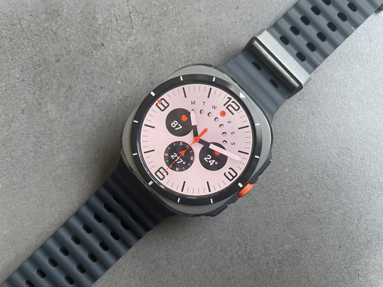 Smartwatch Samsung Galaxy Watch Ultra z czarnym silikonowym paskiem i jasnoróżowym wyświetlaczem, pokazujący godzinę, datę, temperaturę i puls.