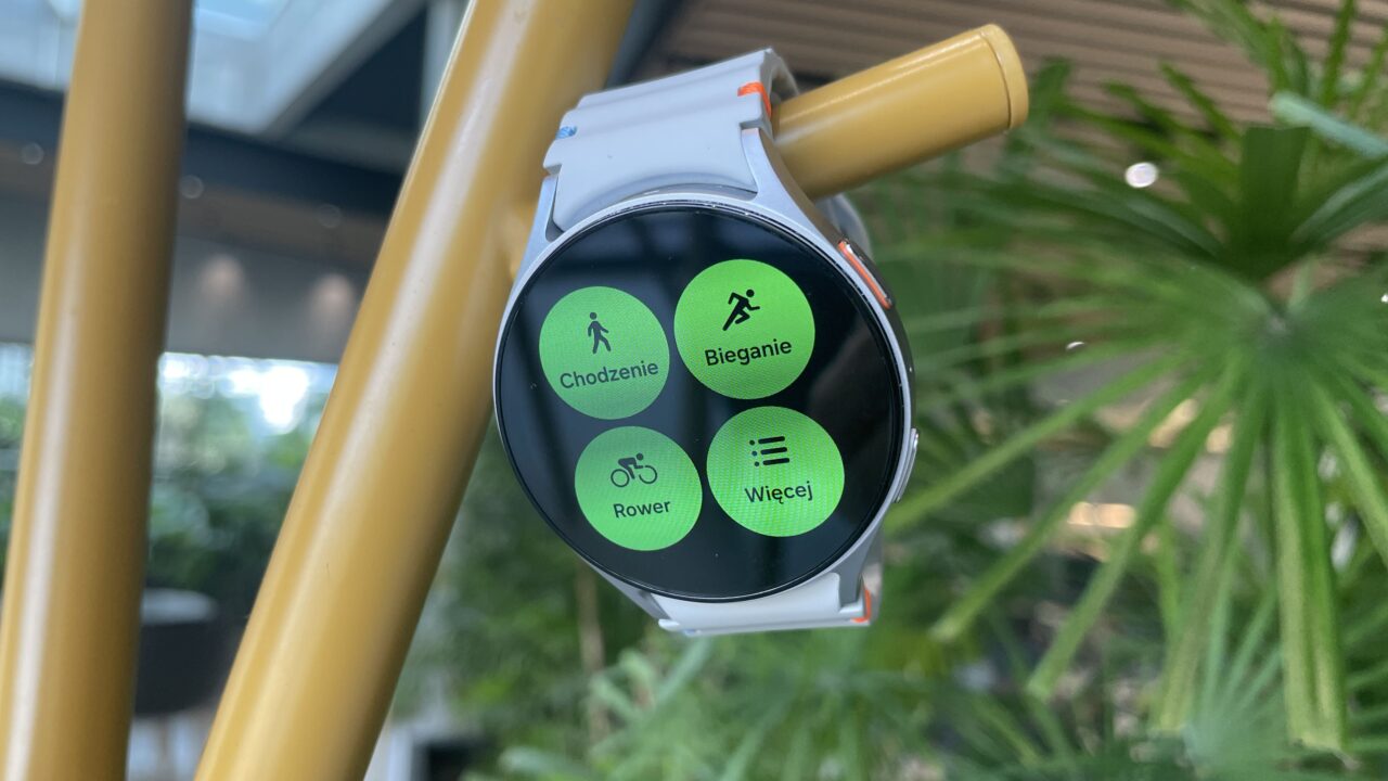 Biały smartwatch z włączonym ekranem, wyświetlającym opcje sportowe: "Chodzenie", "Bieganie", "Rower" i "Więcej".