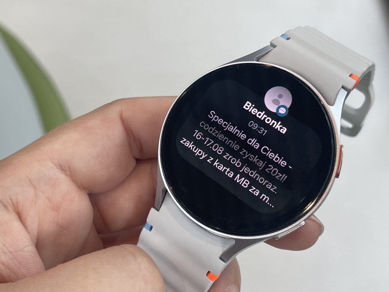 Smartwatch z białym paskiem, wyświetlający wiadomość z aplikacji Biedronka.