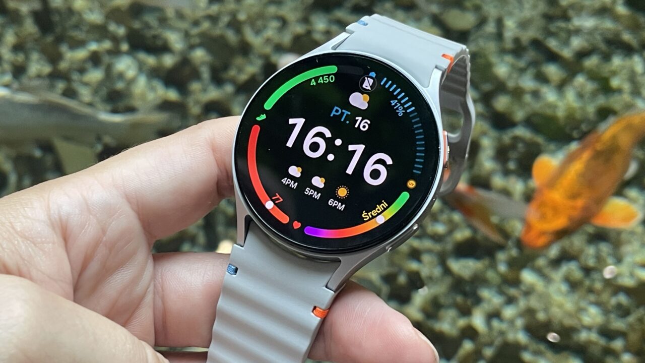 Zegarek smartwatch z szarą opaską, trzymany w dłoni przed akwariem z rybami.