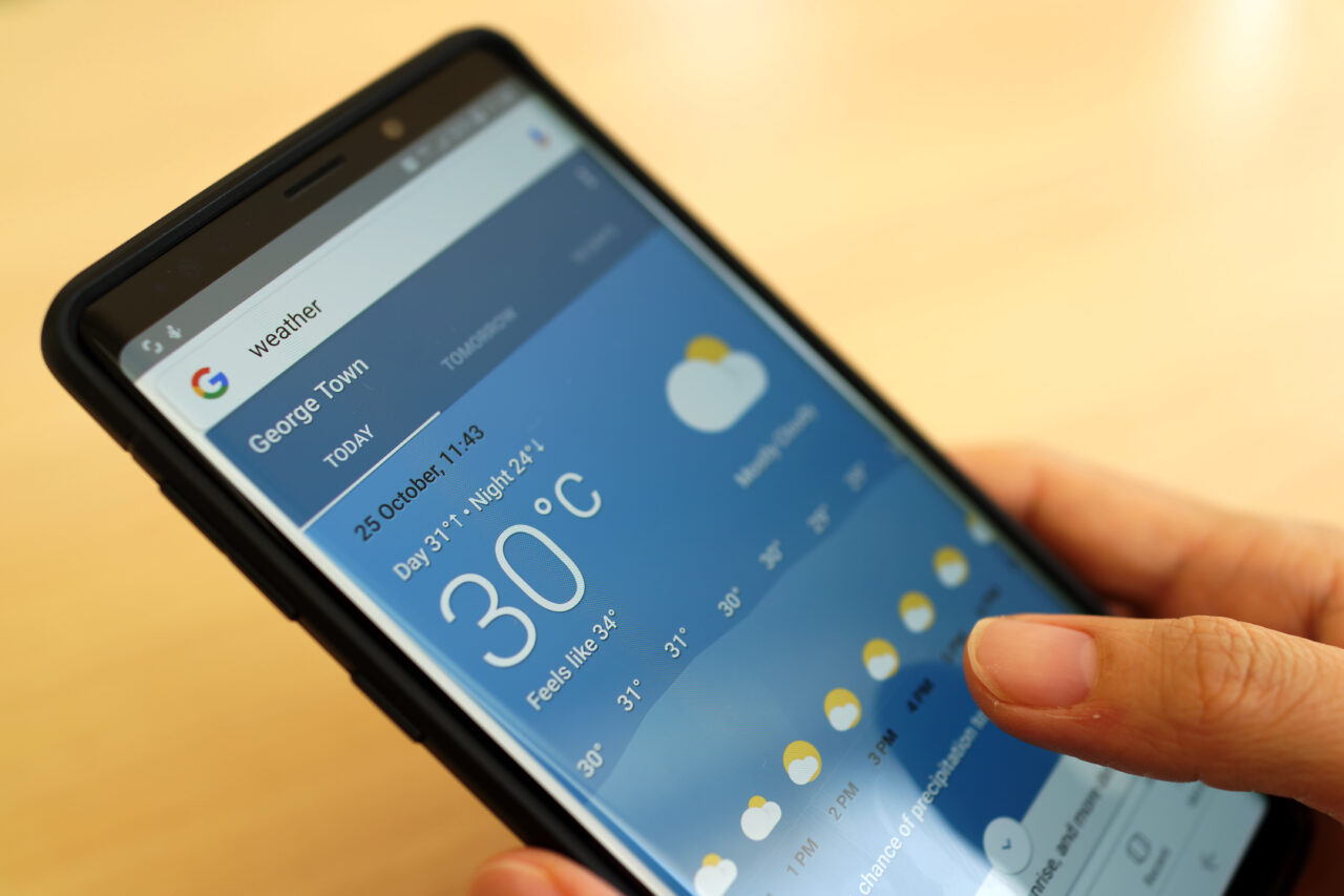 Pogoda od Google Wearther. Osoba trzymająca smartfon z aplikacją pogodową pokazującą temperaturę 30°C w mieście George Town.