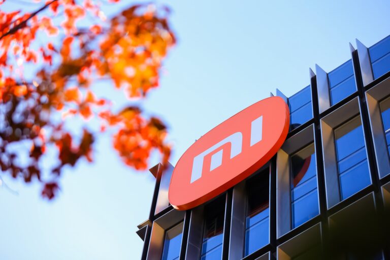 Logo Xiaomi na fasadzie budynku z widokiem na gałęzie drzewa z czerwonymi liśćmi na pierwszym planie.