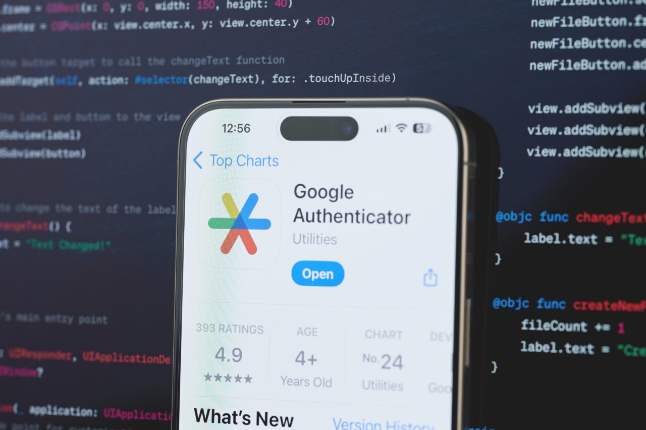 Telefon z aplikacją Google Authenticator na ekranie, w tle kod programistyczny.