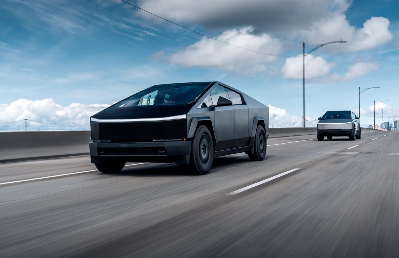 Tesla Cybertruck. Dwa futurystyczne samochody na autostradzie, jasne niebo w tle.