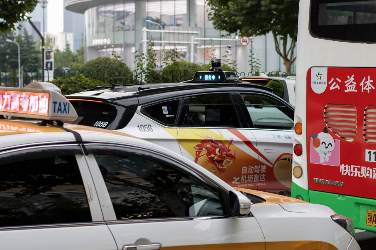 Robotaxi w Wuhan. Autonomiczna taksówka z napisem "Airport Direct Service" obok tradycyjnej żółto-białej taksówki i autobusu na ulicy w mieście.