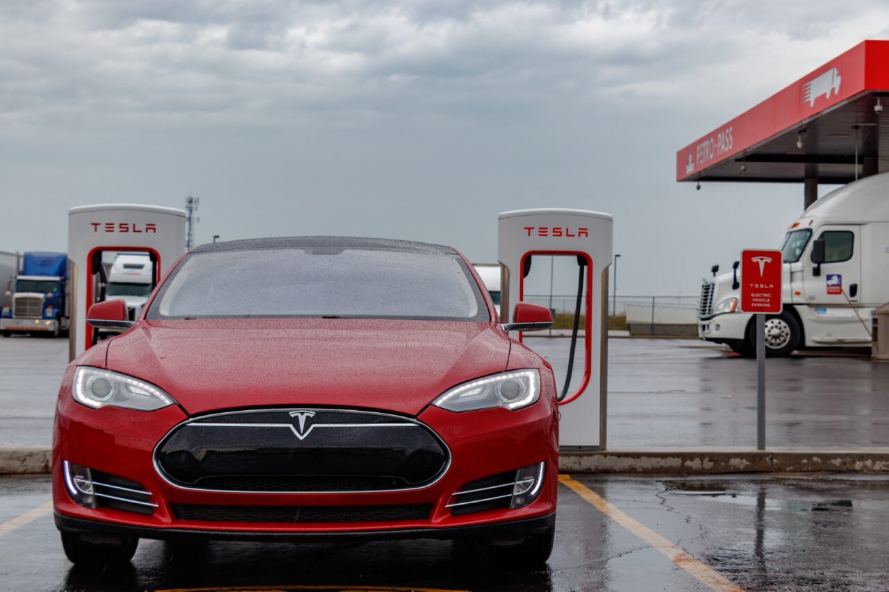 Czerwony samochód Tesla ładujący się na stacji ładowania, w tle parking dla ciężarówek.