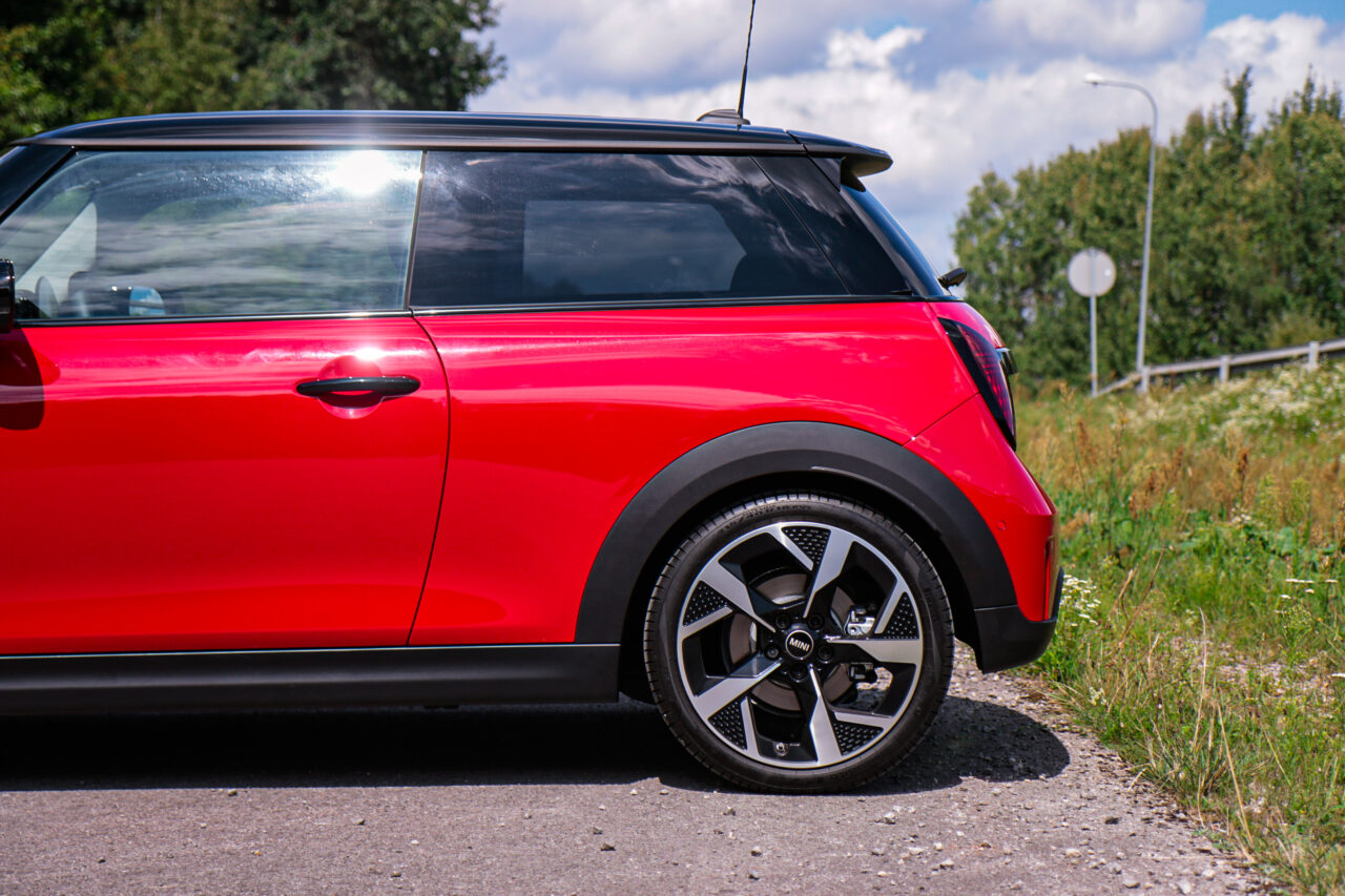 Test Mini Cooper C, czerwony samochód z czarnym dachem na drodze obok zielonych krzewów.