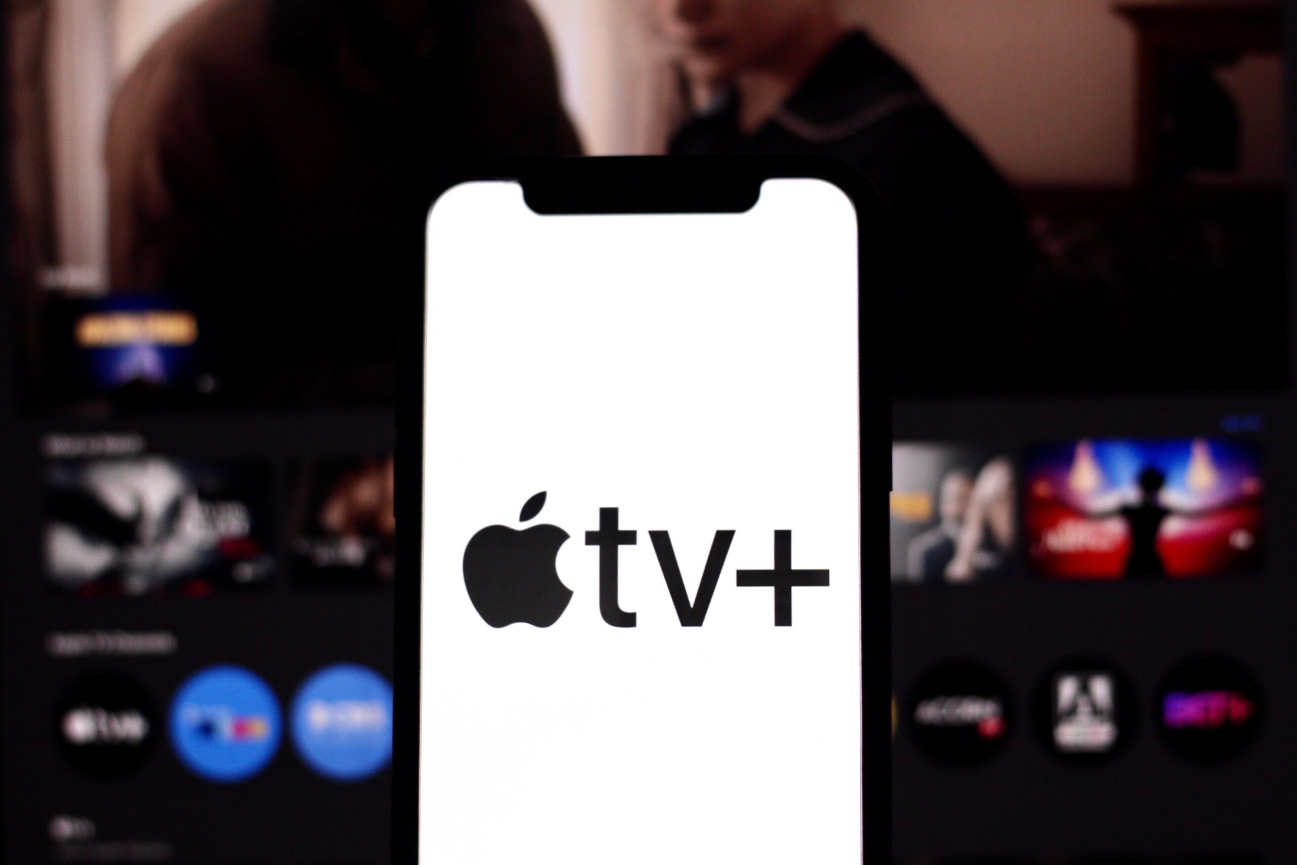 Logo Apple TV+ na ekranie smartfona na tle rozmytych obrazów telewizyjnych.