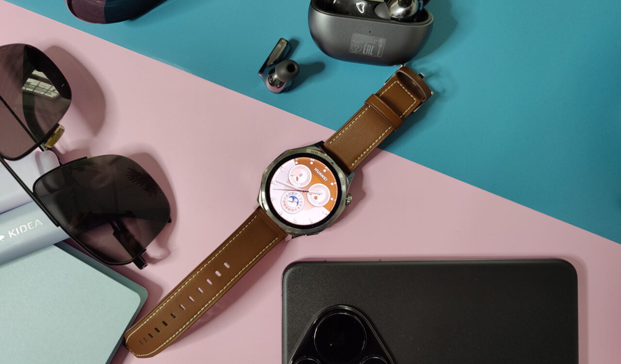 Smartwatch HUAWEI WATCH GT 5 z brązowym skórzanym paskiem na kolorowym tle, otoczony okularami przeciwsłonecznymi, słuchawkami i innymi przedmiotami.