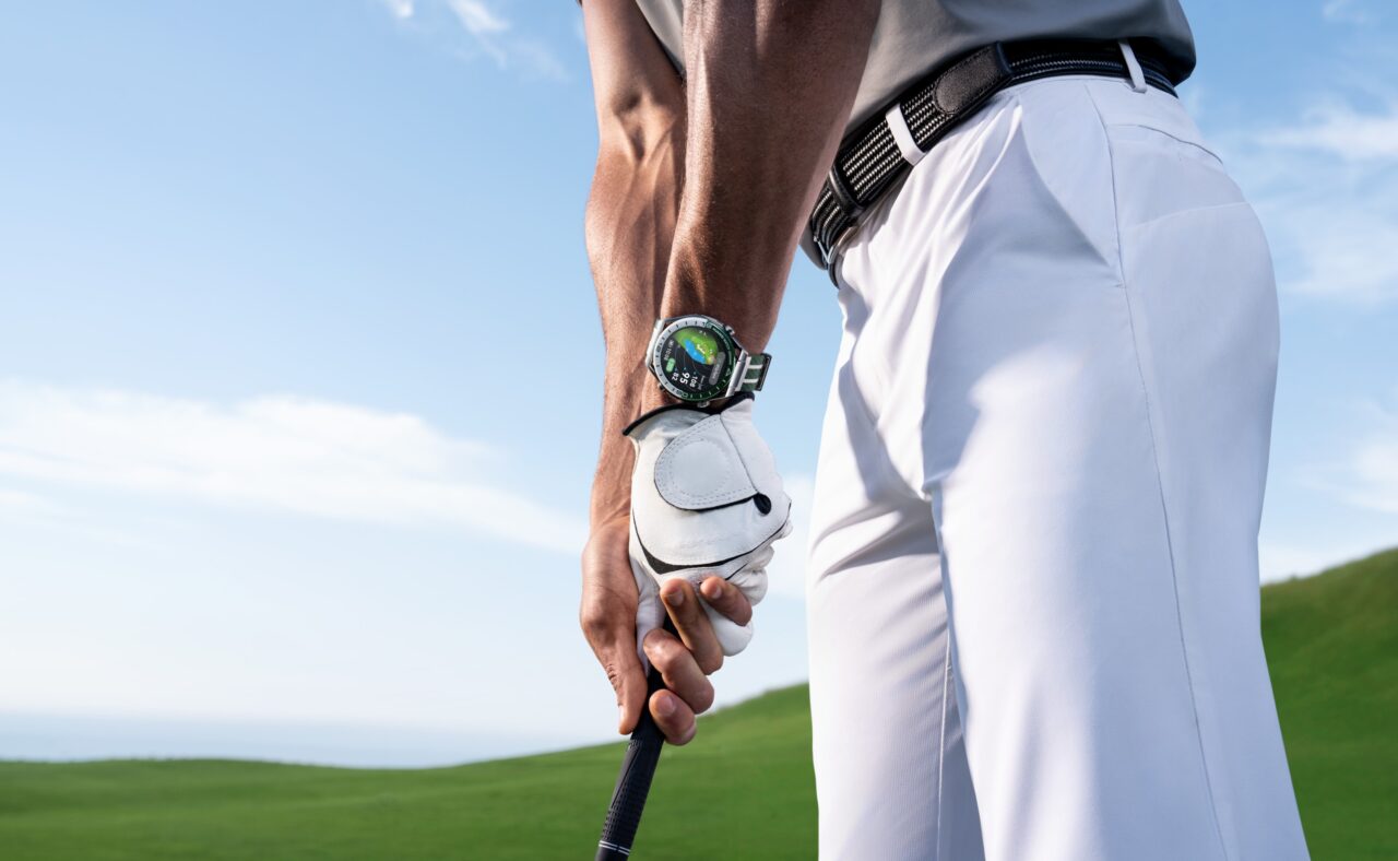 Huawei Watch Ultimate Green - Osoba ubrana w biały golfowy strój trzymająca kij golfowy, na lewej ręce ma zegarek sportowy.