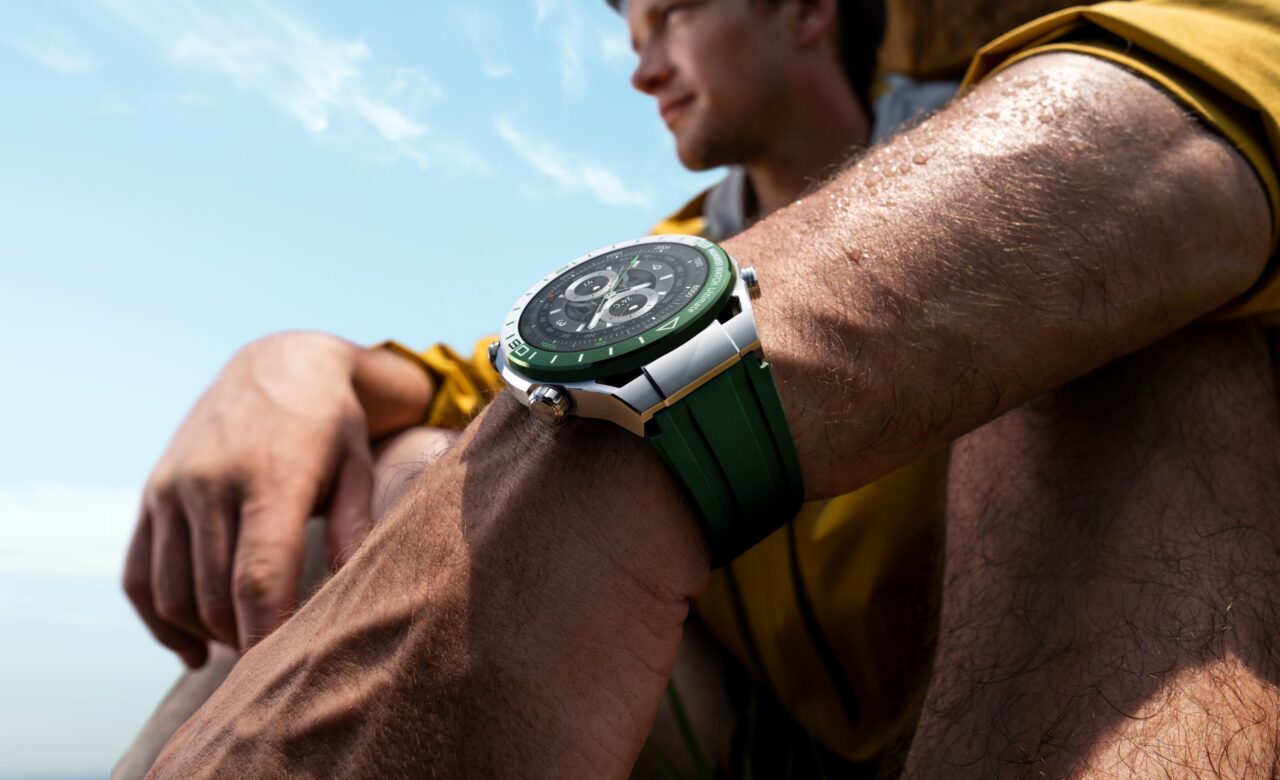 HUAWEI WATCH Ultimate Green - Mężczyzna w żółtej kurtce siedzi, nosząc zielony zegarek sportowy z metalową kopertą na nadgarstku, na tle niebieskiego nieba.