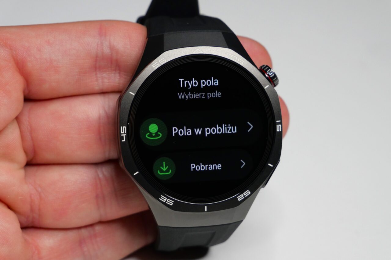 Zbliżenie na smartwatch z wyświetlaczem w języku polskim, trzymany w ręku, pokazujący opcje menu: "Tryb pola", "Pola w pobliżu" i "Pobrane".