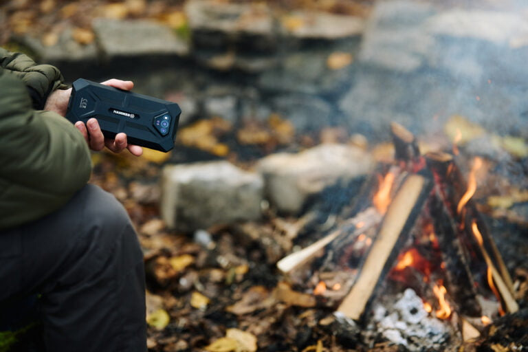 Osoba trzymająca wytrzymały smartfon marki Hammer, siedząca w pobliżu ogniska w lesie.