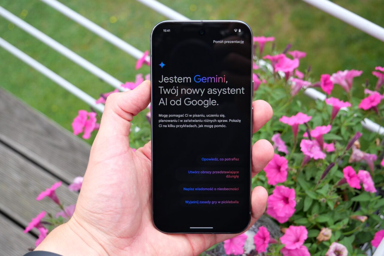 Osoba trzymająca telefon z aplikacją Gemini na ekranie. W tle kwiaty.