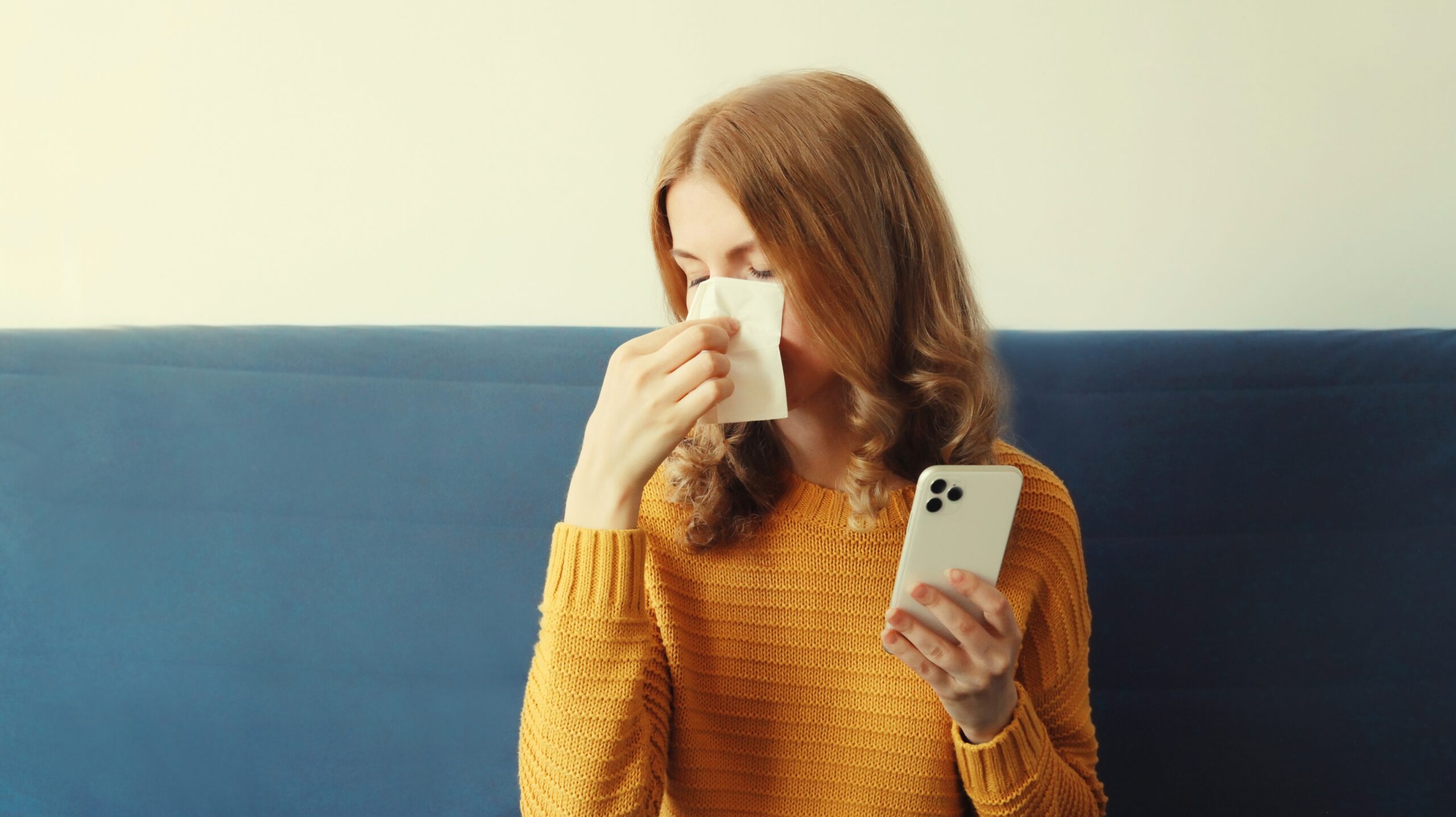 Kobieta w żółtym swetrze wycierająca nos chusteczką, trzymająca telefon komórkowy w ręce.