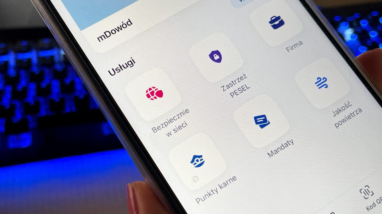 Smartfon z uruchomioną aplikacją mobilną, wyświetlającą różne usługi: bezpiecznie w sieci, zastrzeż PESEL, firma, jakość powietrza, mandaty, punkty karne.