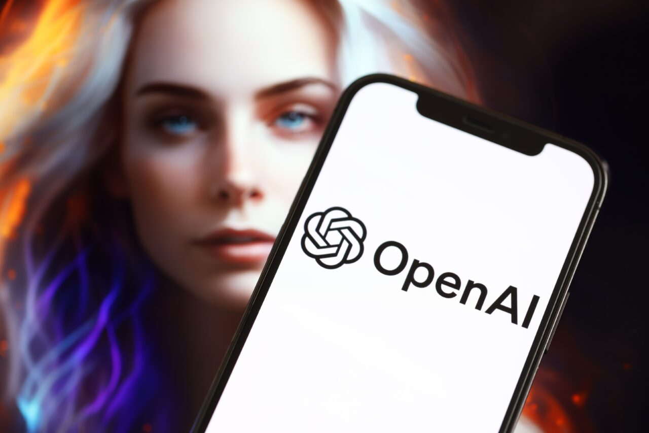 Logo OpenAI na ekranie smartfona, za którym widać rozmyty obraz kobiety z jasnymi włosami.