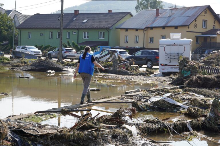 Osoba w kamizelce idzie obok zniszczeń po powodzi; w tle zniszczone samochody i domy.