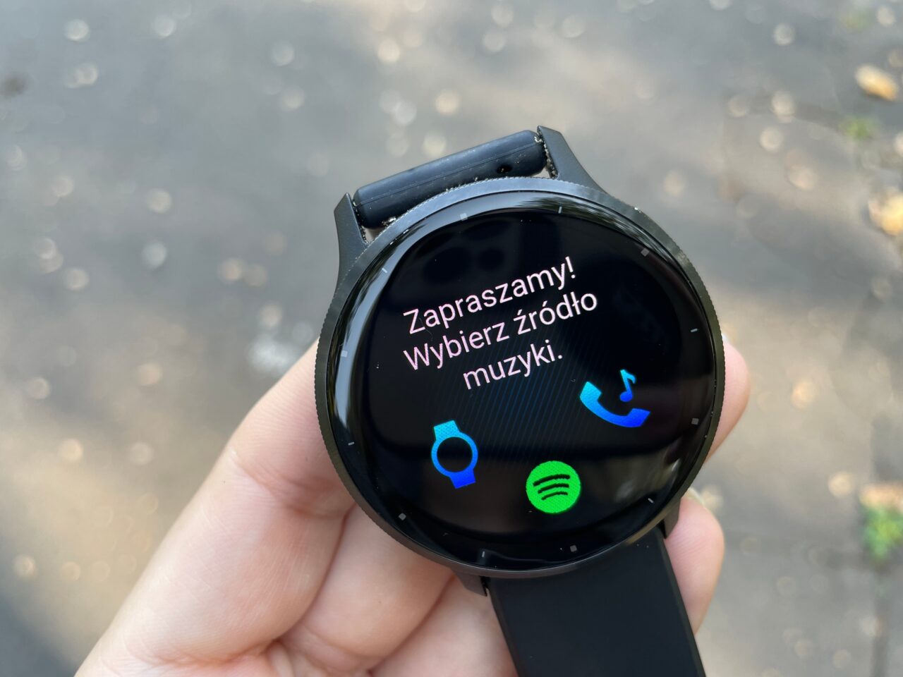 Smartwatch z ekranem wyświetlającym komunikat "Zapraszamy! Wybierz źródło muzyki." i ikony aplikacji do wyboru.