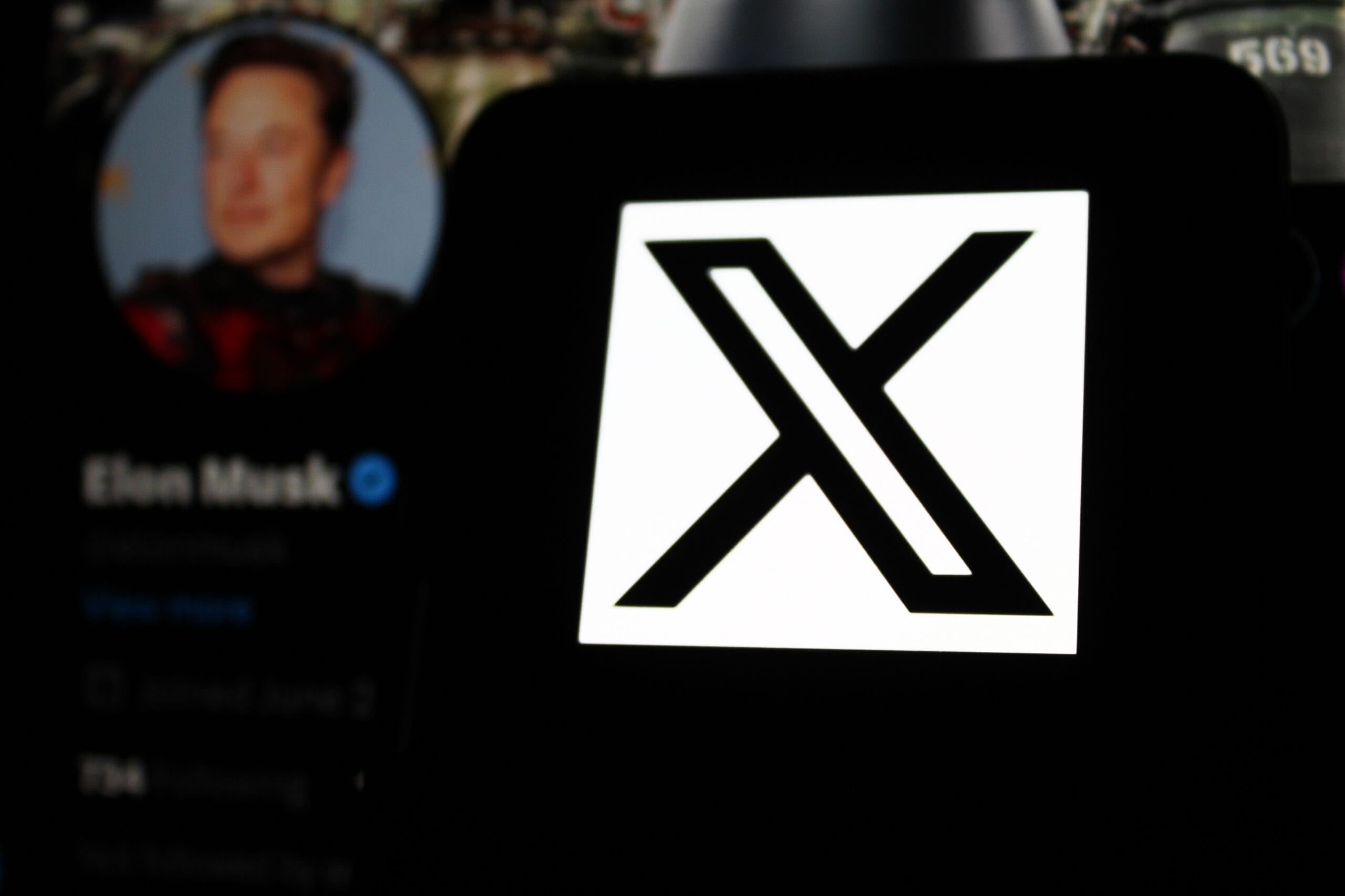 Logo "X" na ekranie telefonu, w tle rozmyty profil użytkownika na Twitterze.