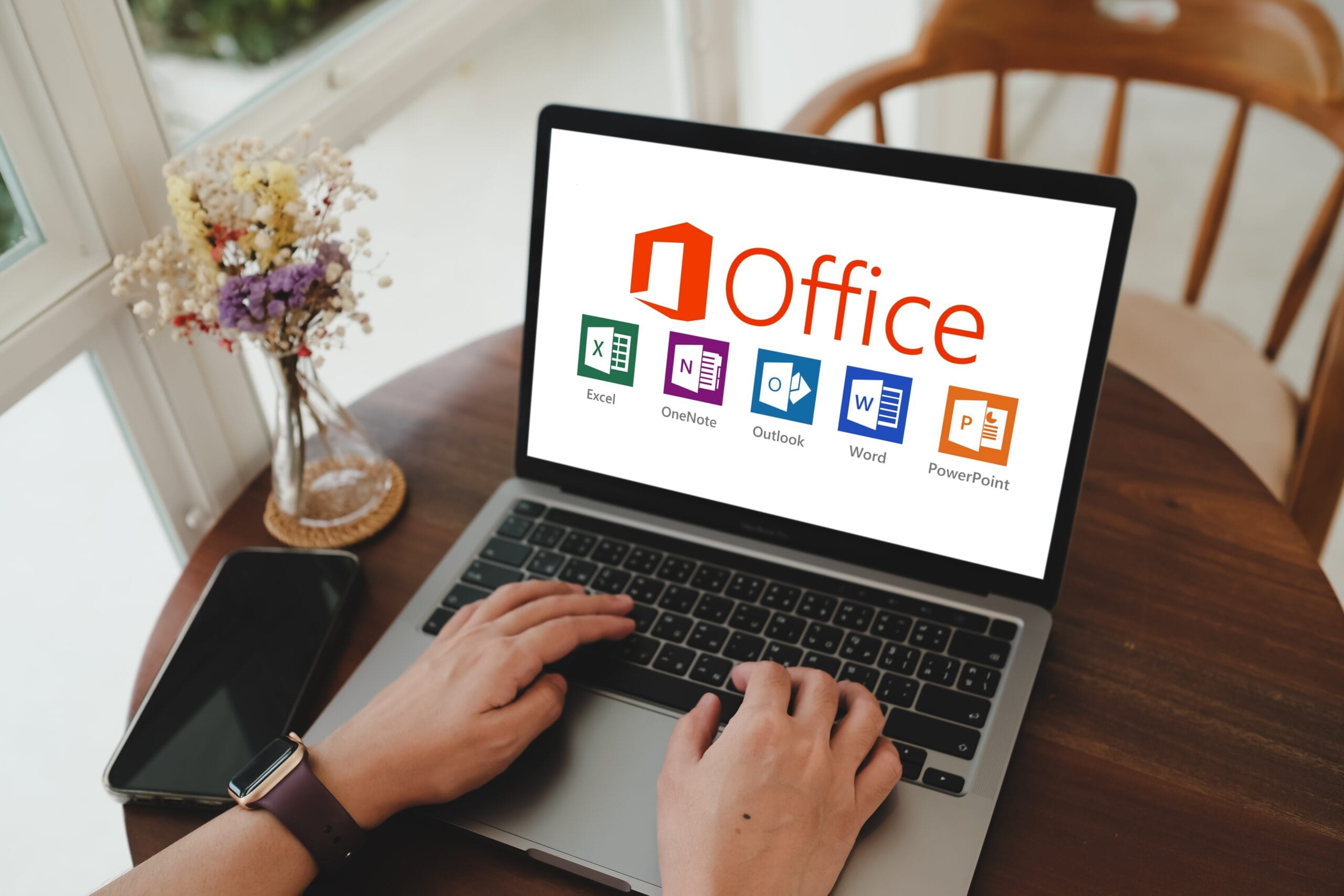 Osoba pisząca na laptopie z wyświetlonym ekranem Microsoft Office, obok leży smartfon, w tle znajduje się wazon z kwiatami.
